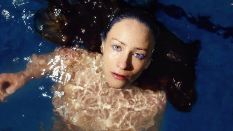 MALLA vedessä kelluvassa muotokuvassa, katse kamerassa, sininen valaistus ja veden heijastukset korostavat elektronisen musiikin tunnelmaa. Kuva: Helmi Padatsu / Meri Björn